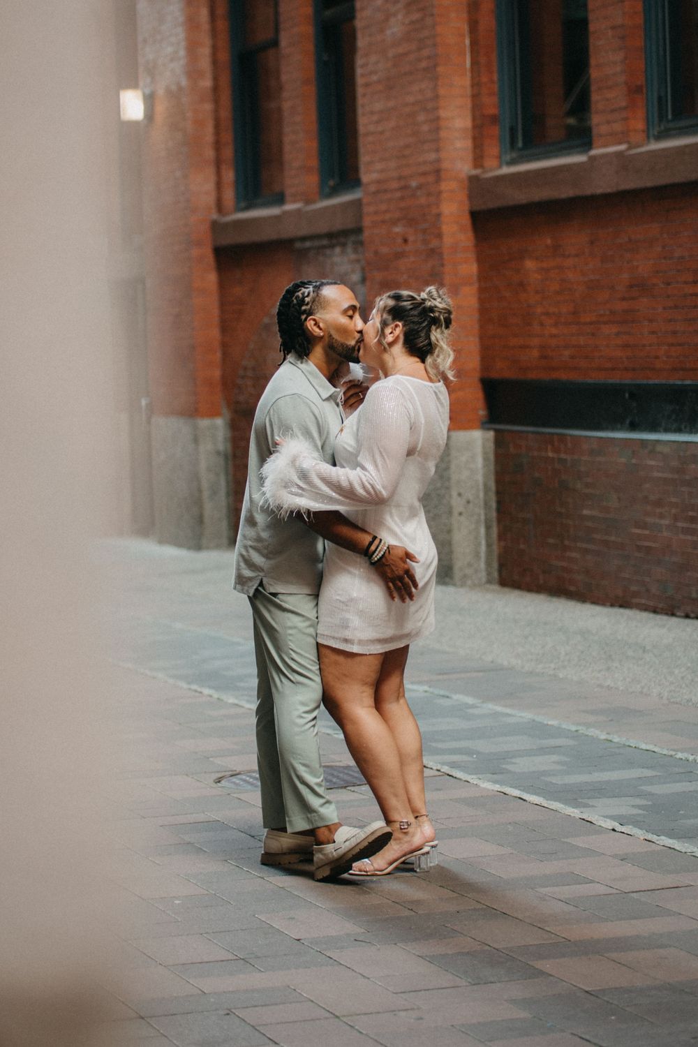 A couple shares a romantic kiss on a city sidewalk, dressed in coordinating neutral tones.