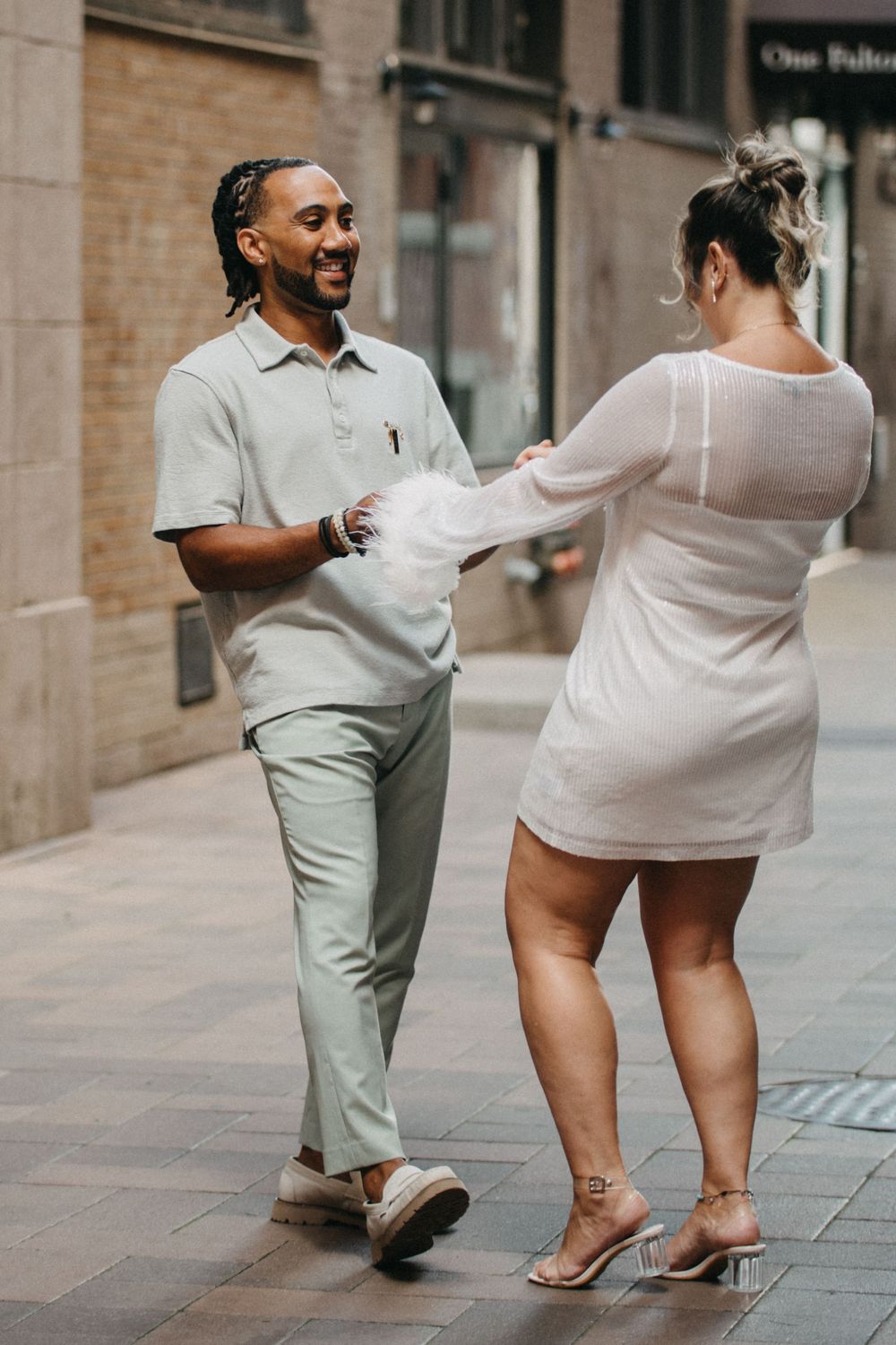 A smiling man in sage green pants holds hands with a woman in a white mini dress on a city street.