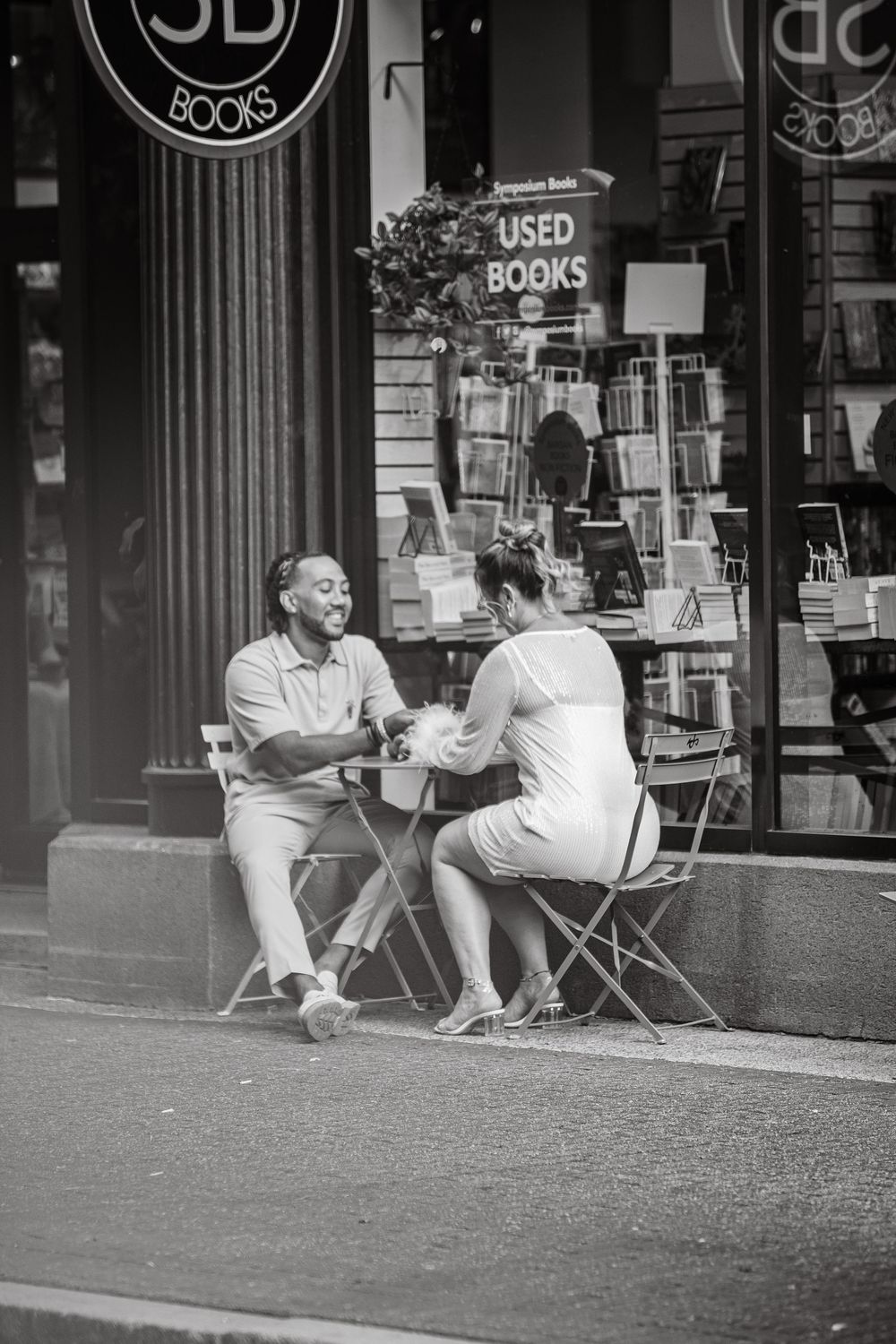 A couple sits and laughs together at an outdoor cafe table in front of a used bookstore.