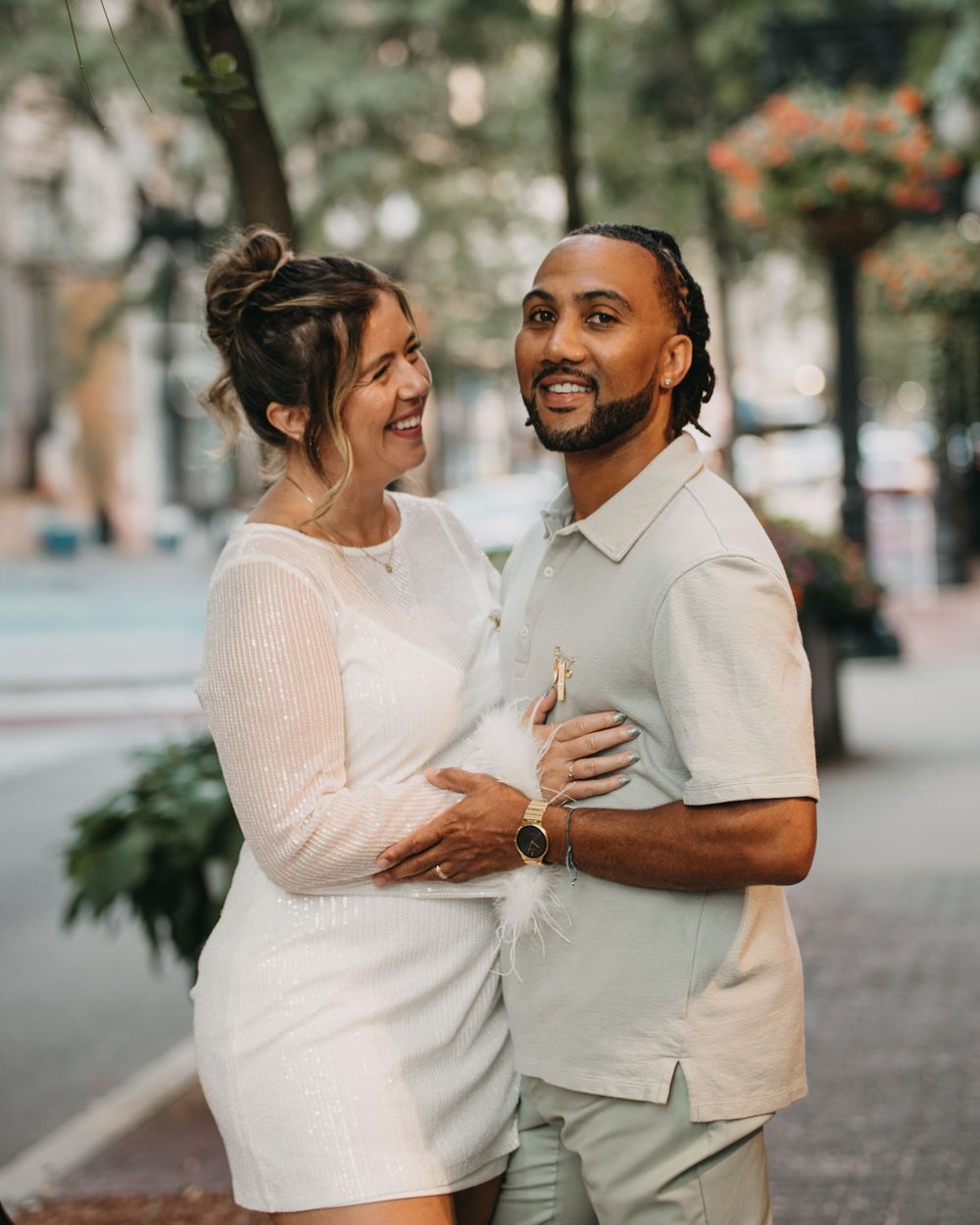 Smiling couple in coordinating white and sage outfits embracing on a tree-lined urban street during engagement photos.