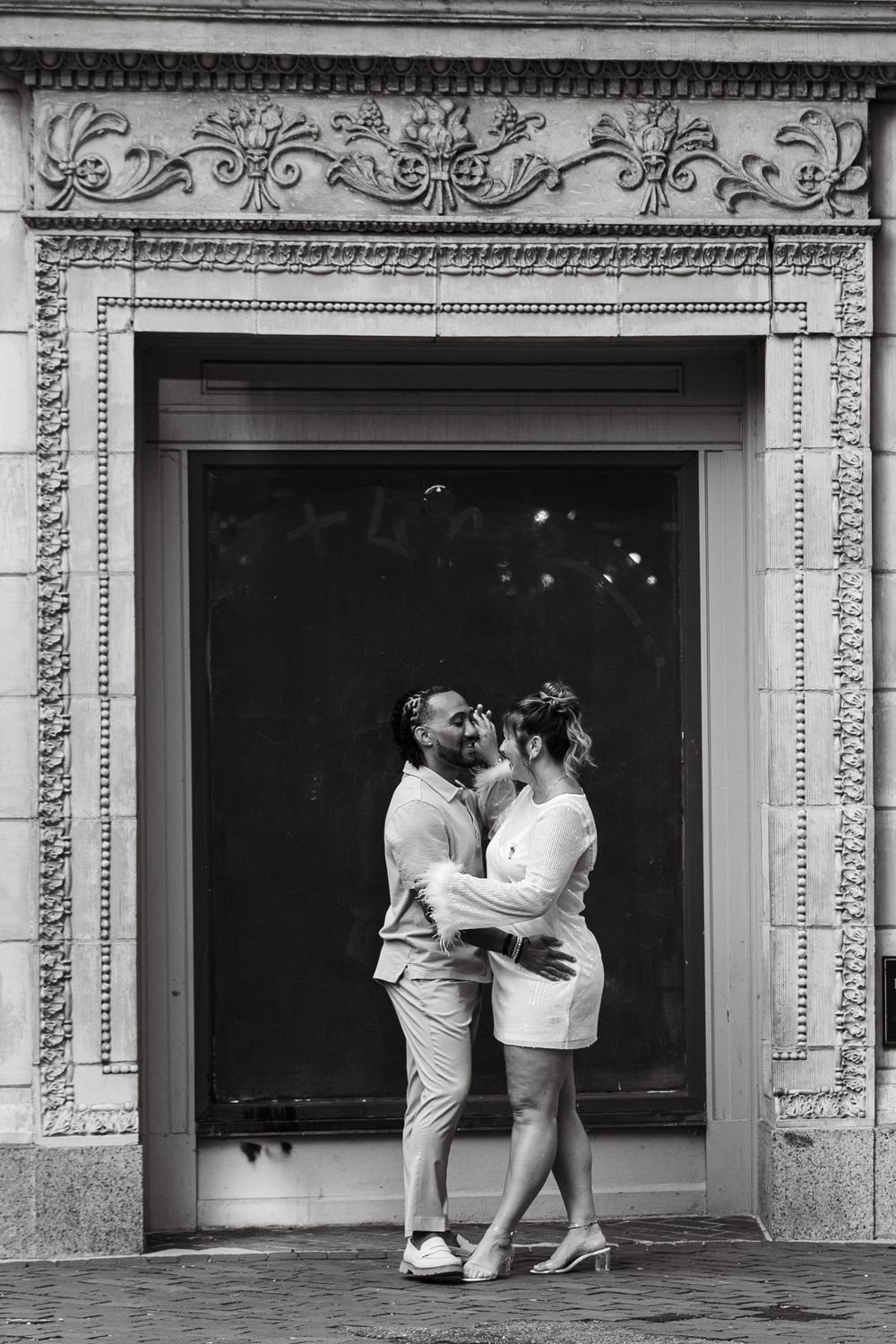 A couple embraces and kisses in front of an ornately carved stone doorway in black and white.
