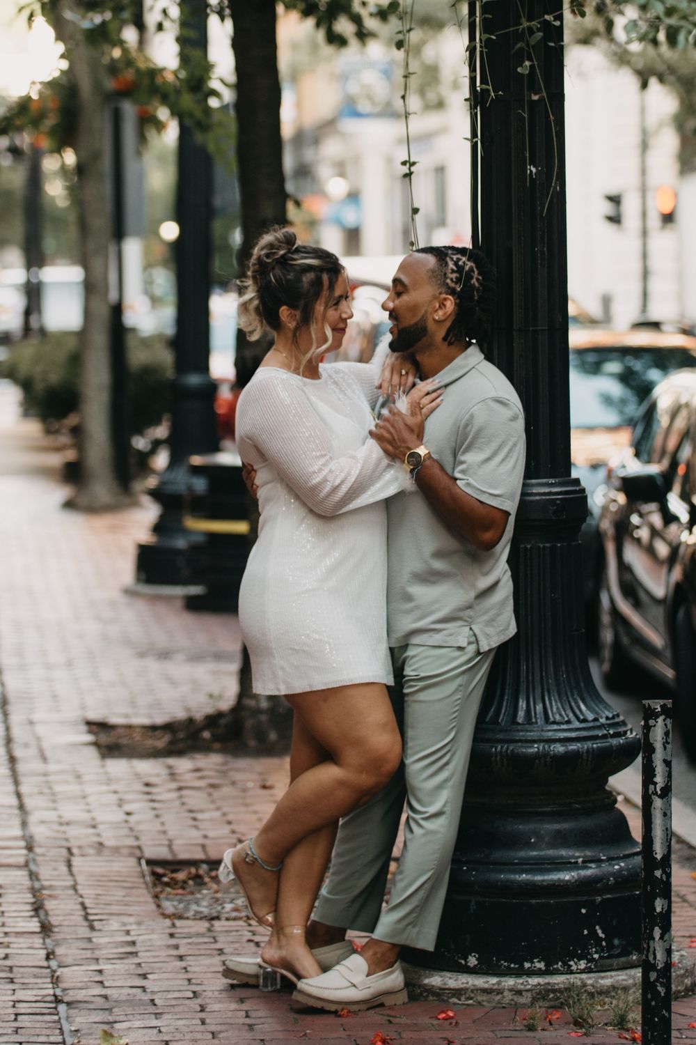 Romantic couple in white and sage outfits leaning against a black lamppost on a brick city sidewalk.