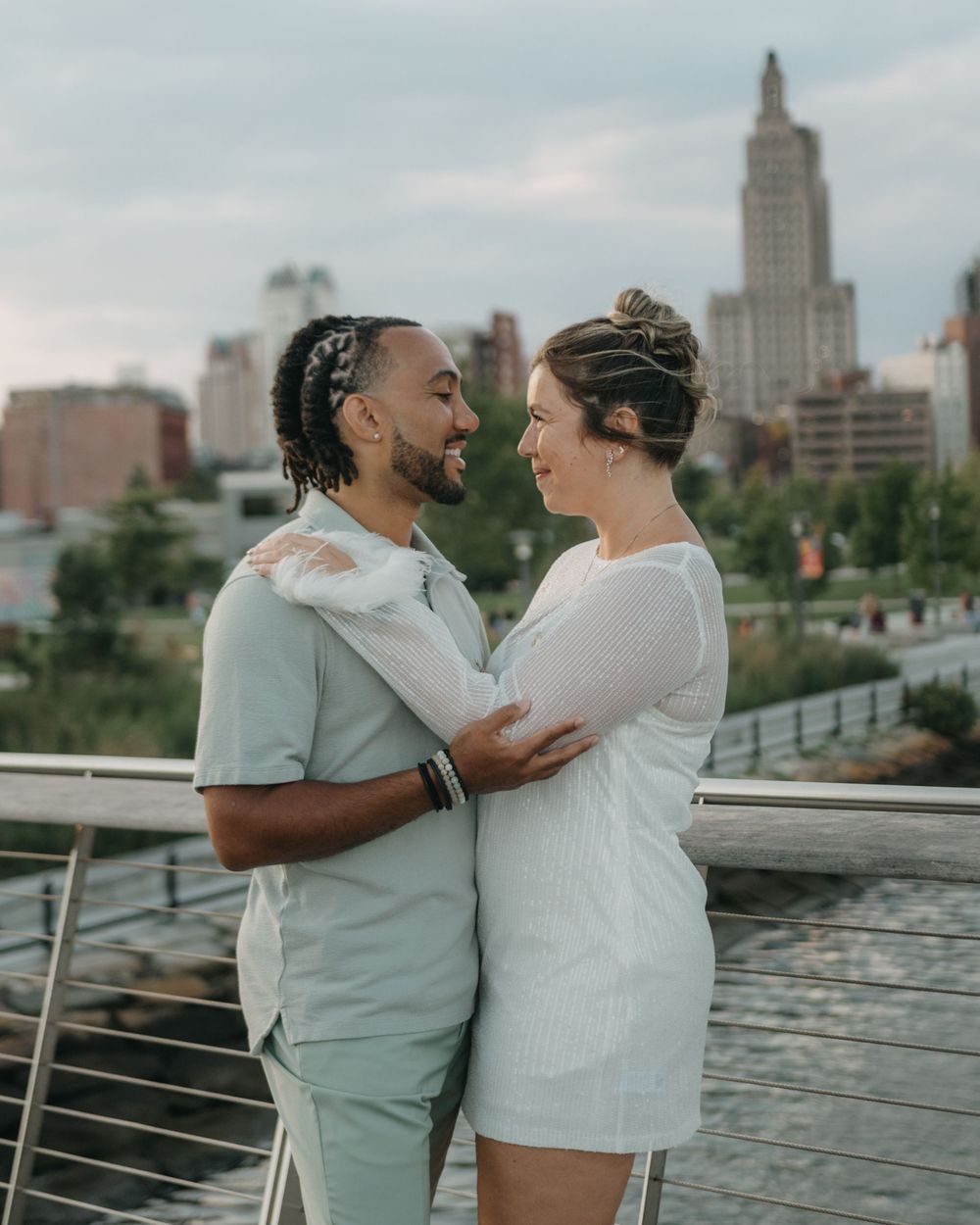 A couple gazes lovingly at each other on a bridge with a city skyline backdrop at dusk.