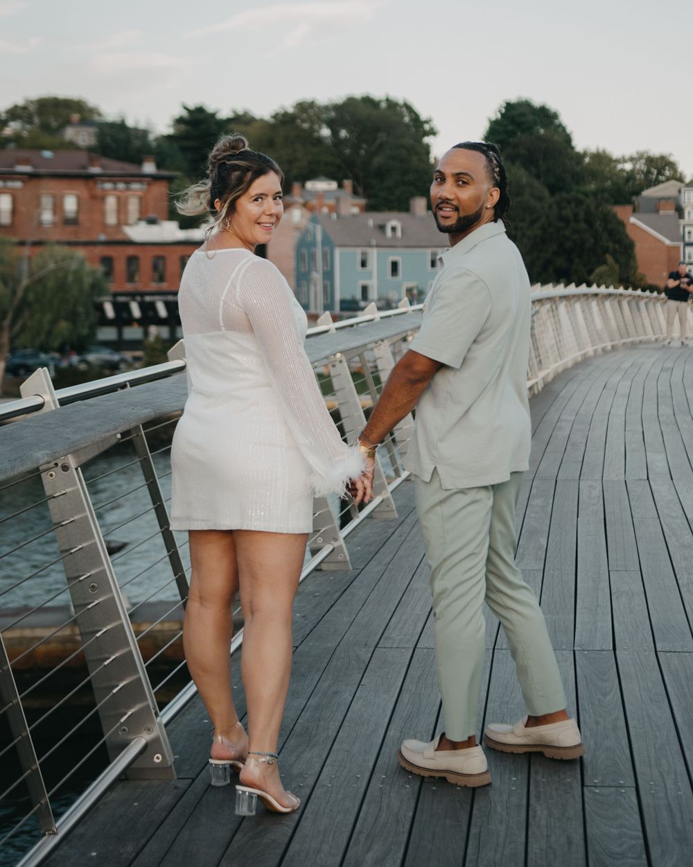 A stylish couple holds hands and looks back while posing on a waterfront boardwalk at sunset.