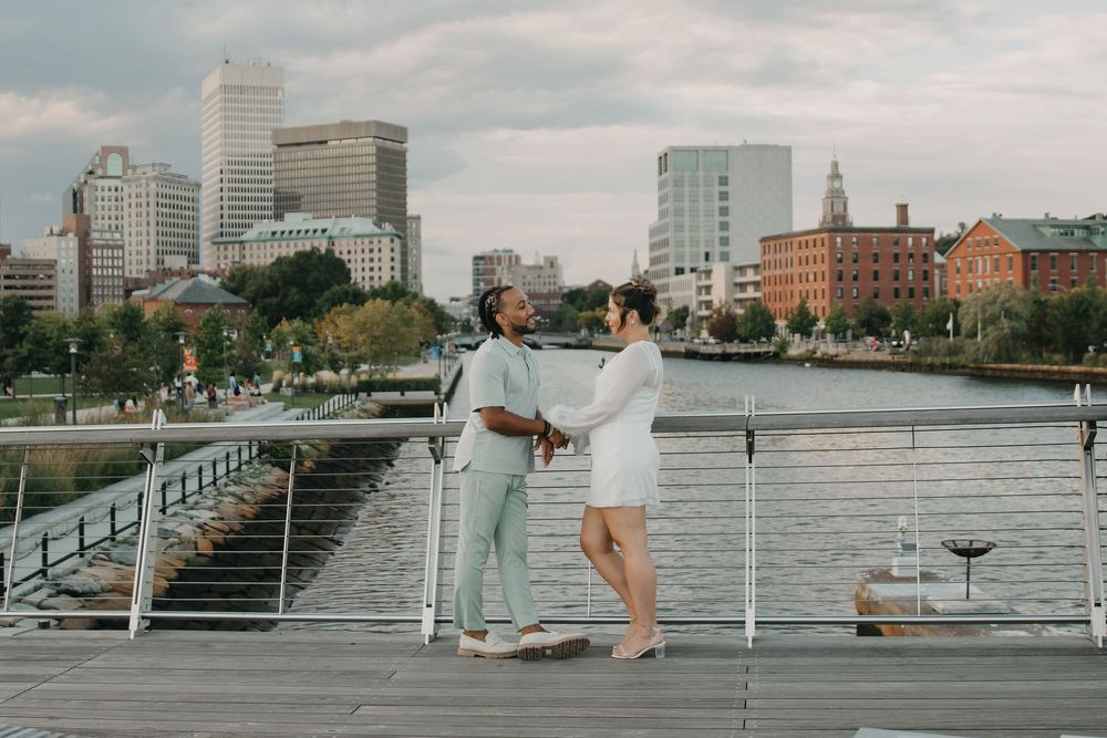 A couple holds hands and smiles on a waterfront bridge with a scenic city skyline behind them.