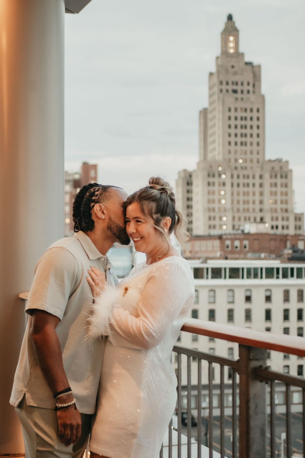 Couple sharing a tender moment on a rooftop balcony with a city skyline and tall building at dusk.