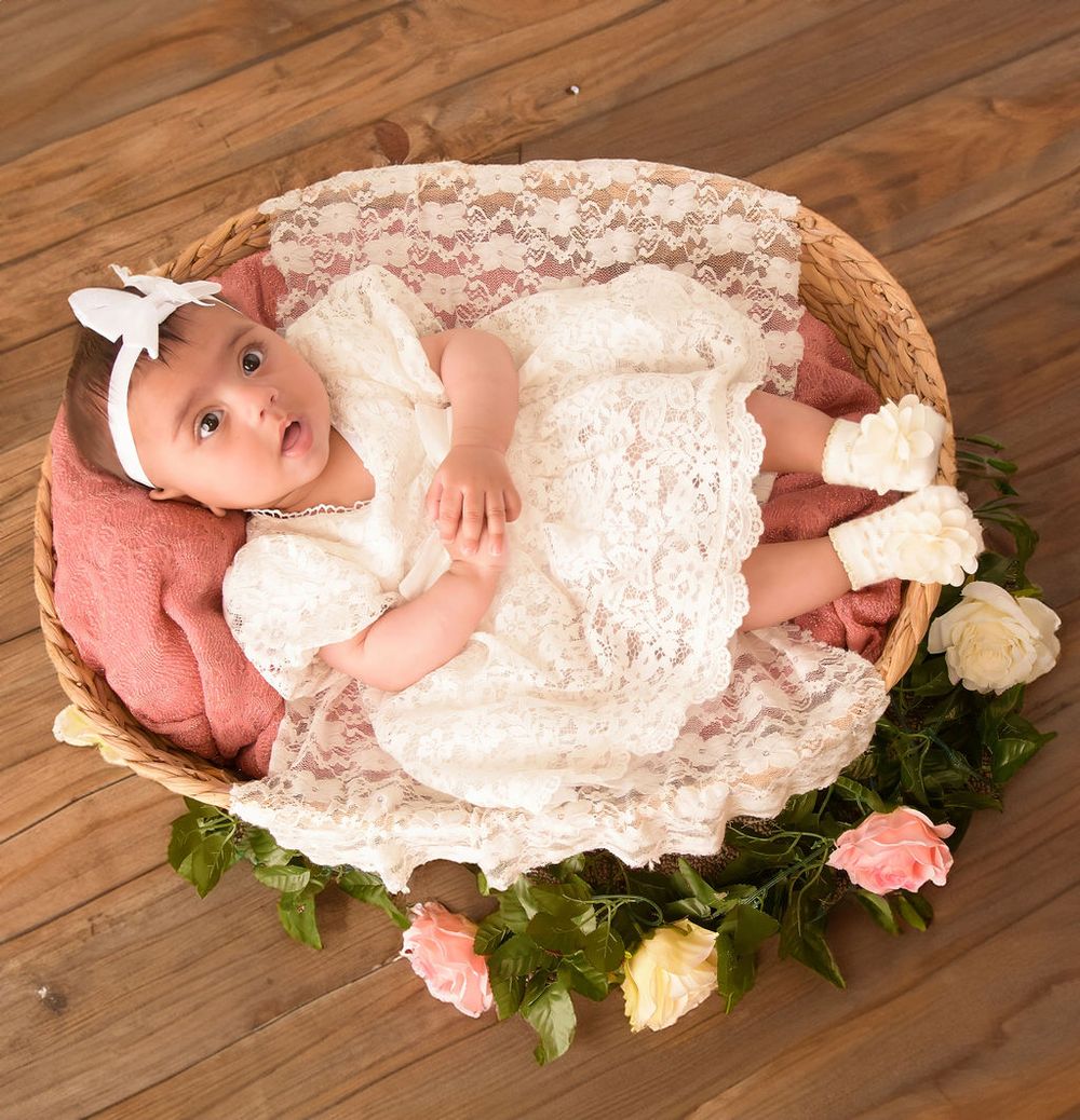 Infant in lace dress posed in basket with roses on wooden floor.