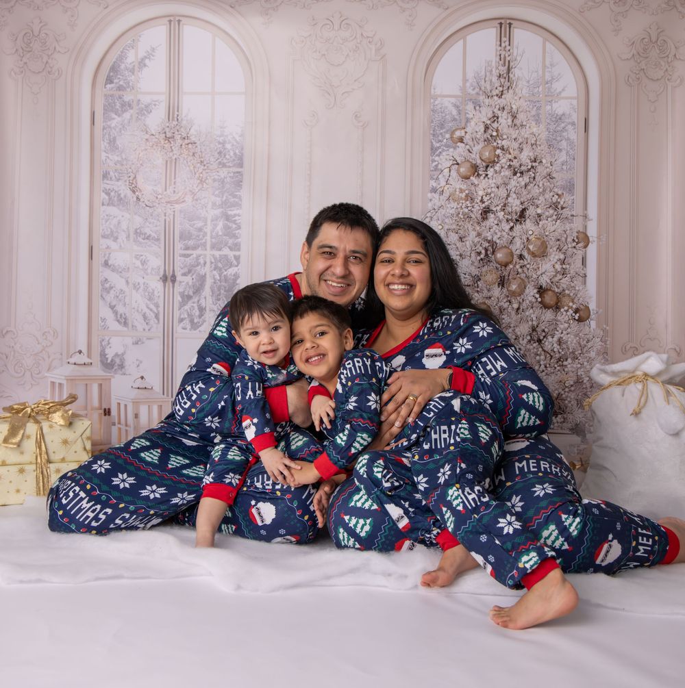 Family wearing matching navy blue holiday pajamas poses together on white floor with Christmas decor background.
