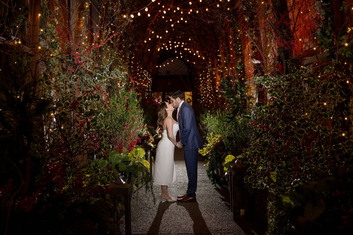 An evening wedding photo in a tunnel decorated with twinkling lights and greenery.