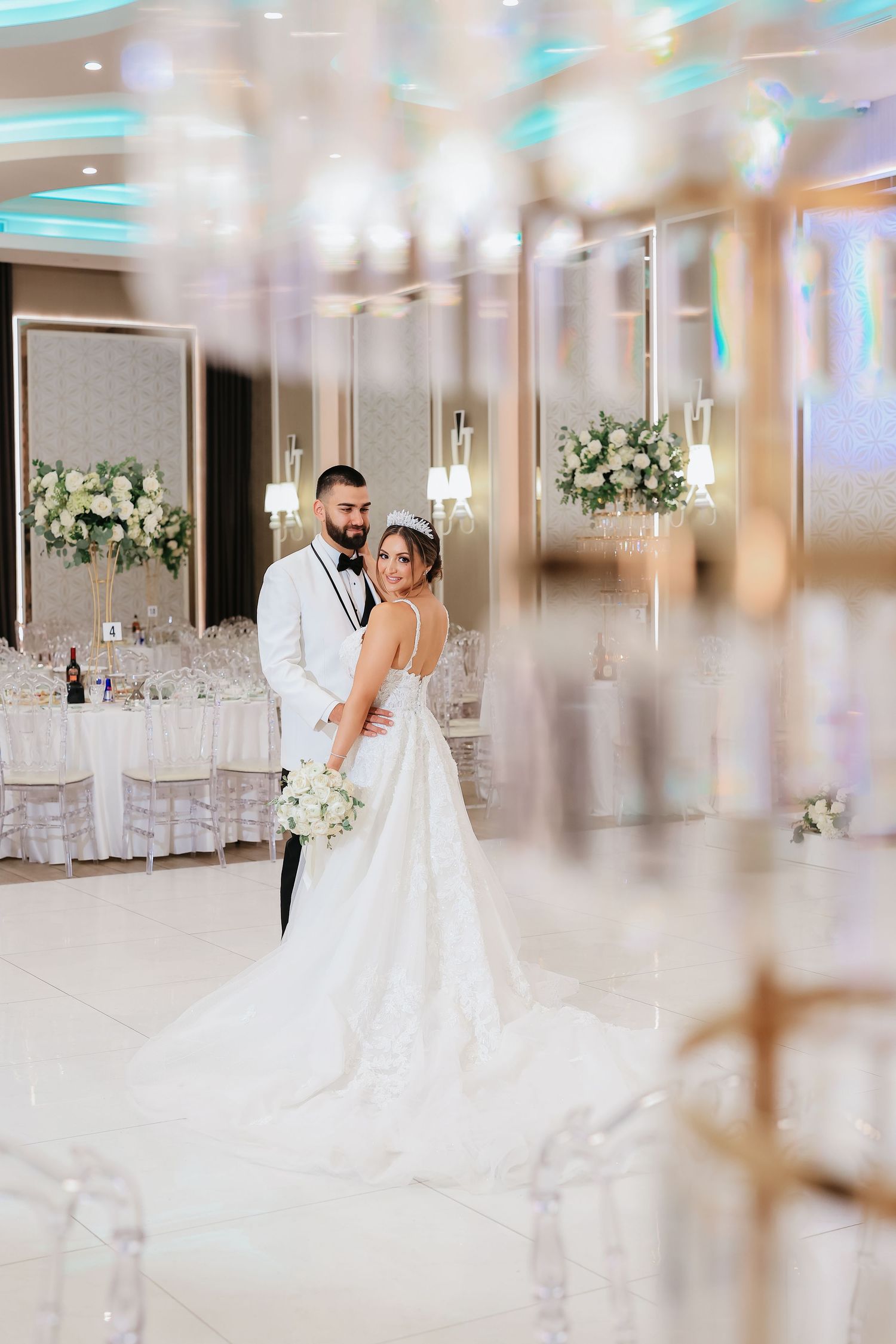 Two people in wedding attire embrace in an elegant ballroom with white floral arrangements and twinkling lights.