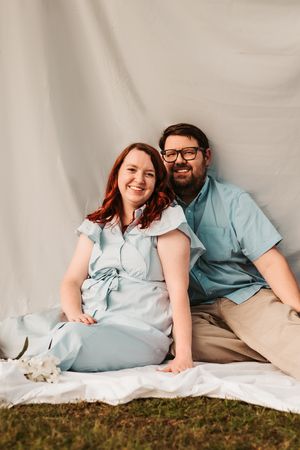 A couple in light blue outfits relaxes on a white blanket outdoors.