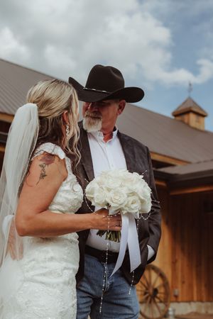 A bride in white dress holds bouquet next to person in cowboy hat outside rustic barn.
