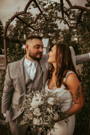 A couple poses with white floral bouquet under a rustic wooden arch.