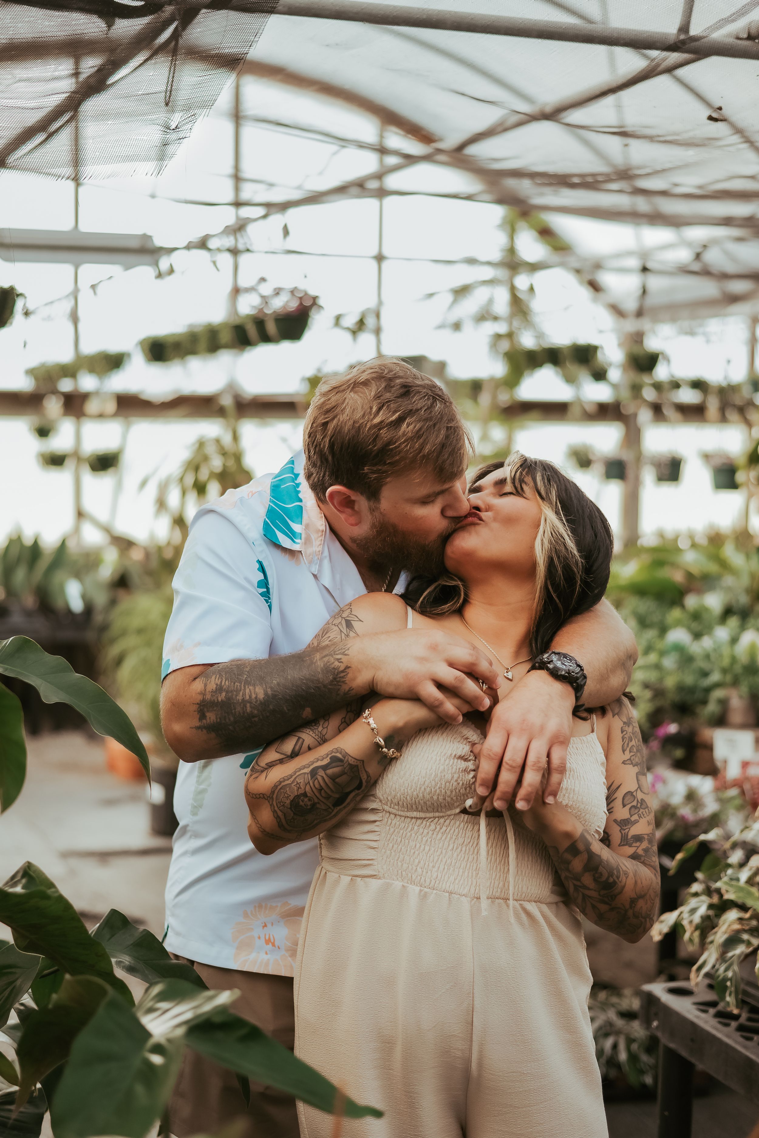 A romantic couple embraces in a greenhouse filled with plants and hanging lights.