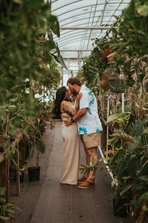 A couple shares an intimate moment in a greenhouse walkway surrounded by lush plants.