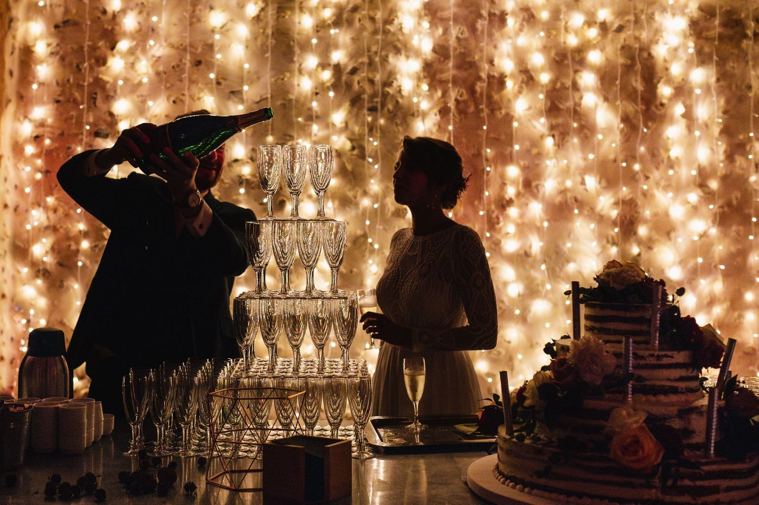 Un couple de jeunes mariés verse le champagne dans une pyramide de coupes. In voit aussi un wedding cake