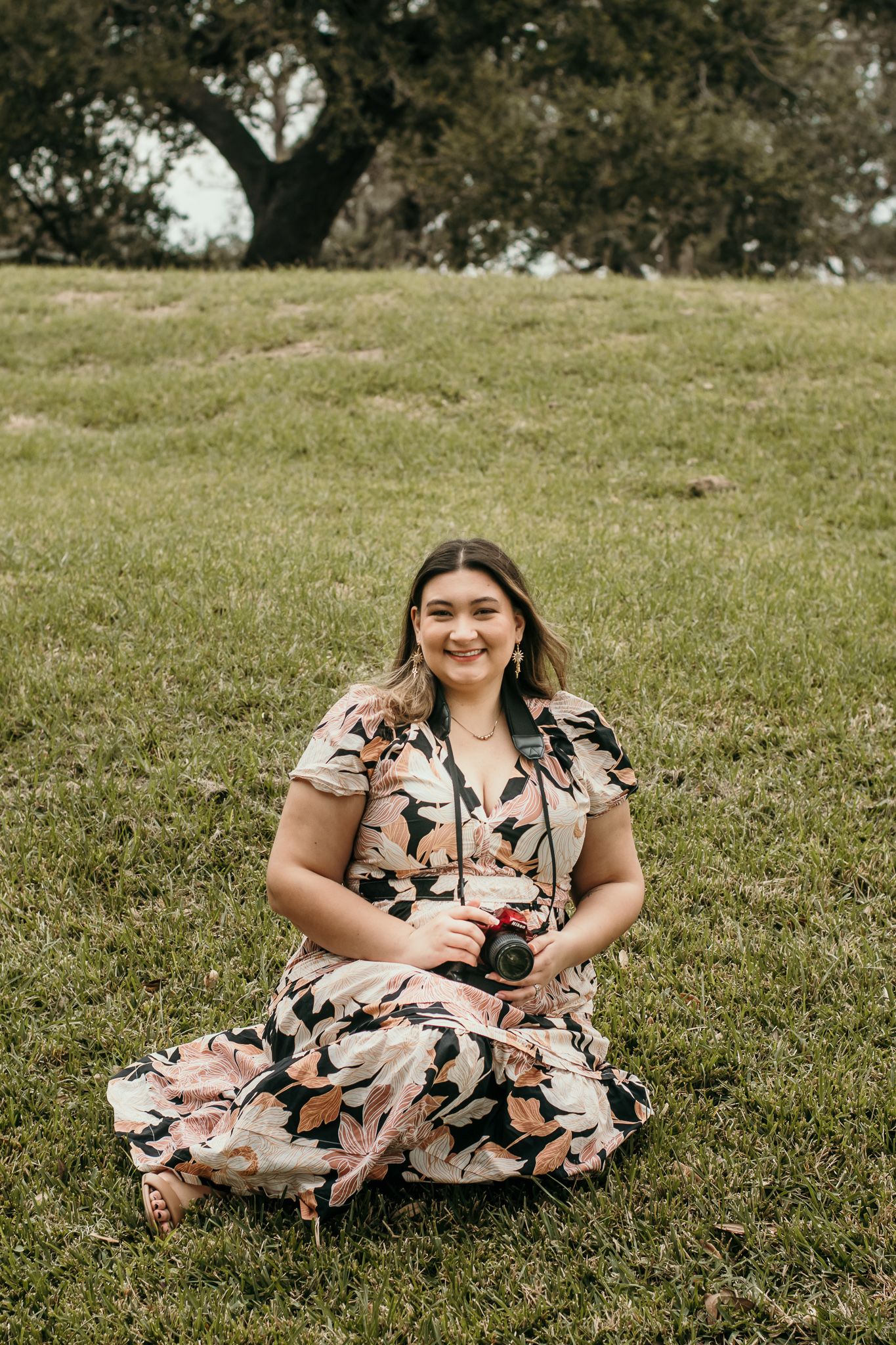 A photographer in a floral dress sits on grassy hill holding a camera on a sunny afternoon.