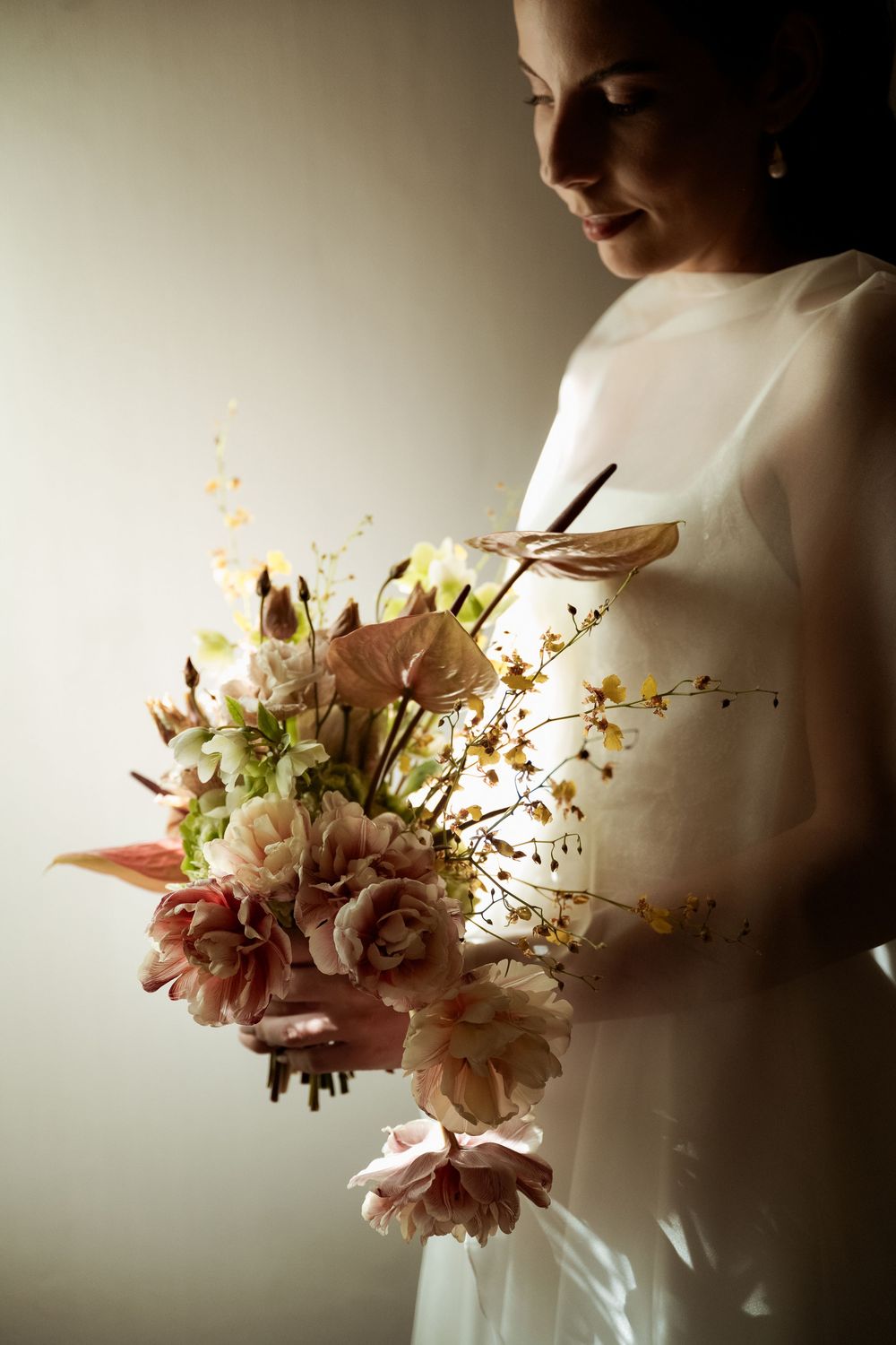 Bride in sheer white gown holds elegant bouquet of blush roses, dahlias, and yellow wildflowers in soft natural light.