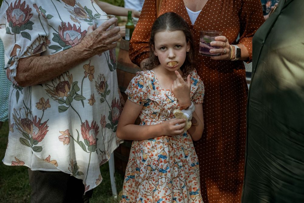 Young girl in floral dress stands outdoors among adults at a garden gathering, looking thoughtful.