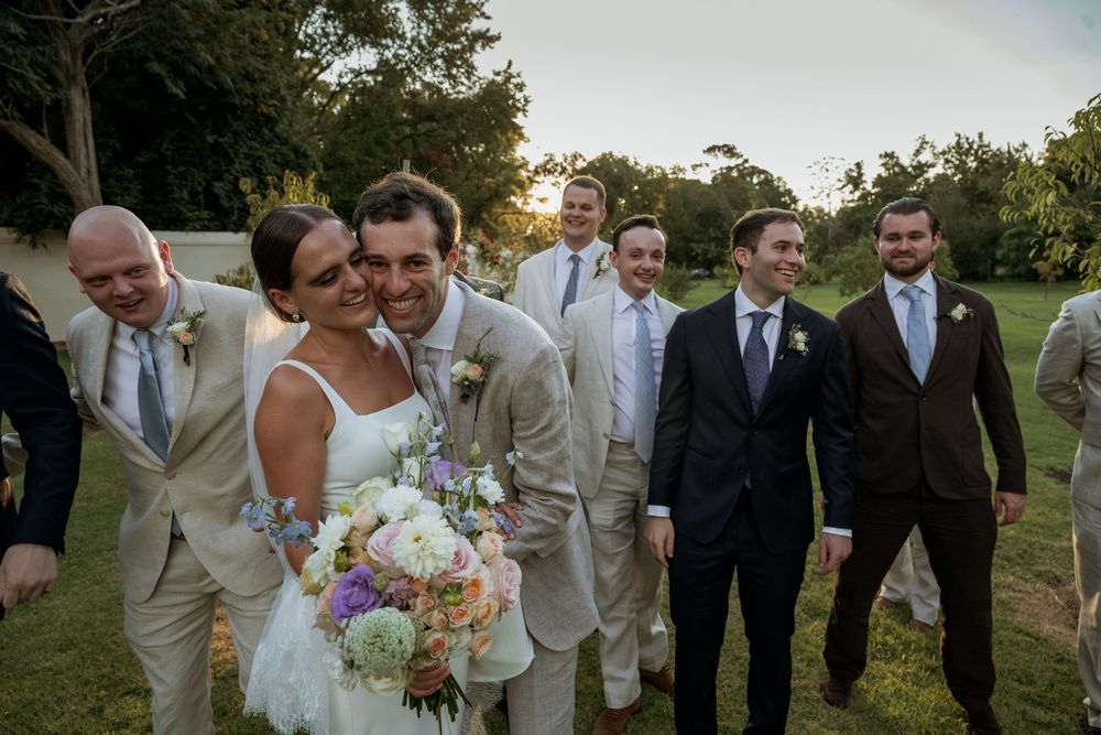 Bride and groom walk with groomsmen in tan suits outdoors at a sunny wedding celebration.
