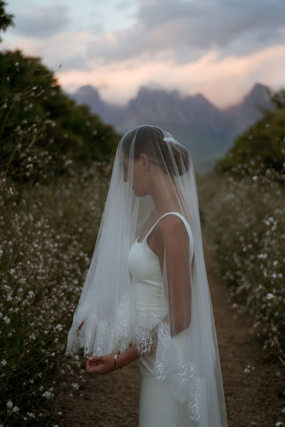Bride in white gown and lace-trimmed veil stands in floral field with misty mountains at dusk.
