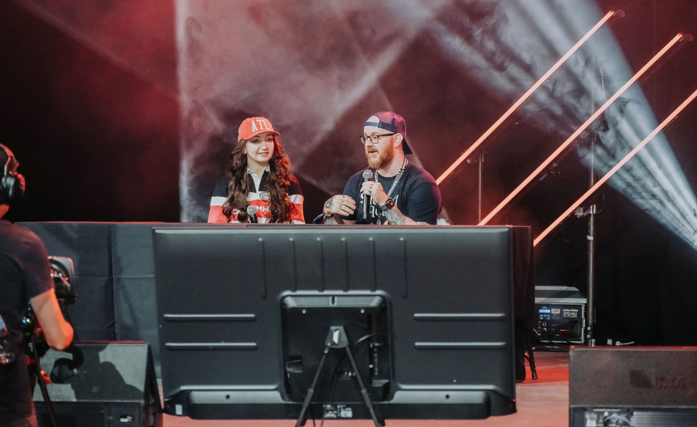 Two commentators at gaming event desk with dramatic lighting and stage effects.