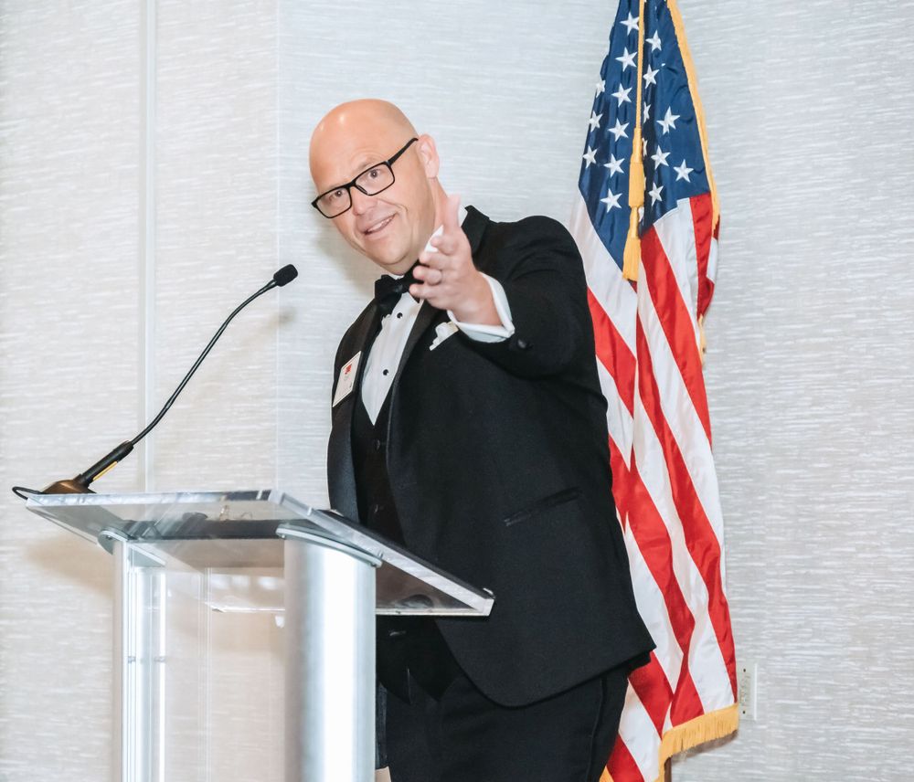 Speaker gestures at podium during presentation with American flag backdrop in formal business attire.