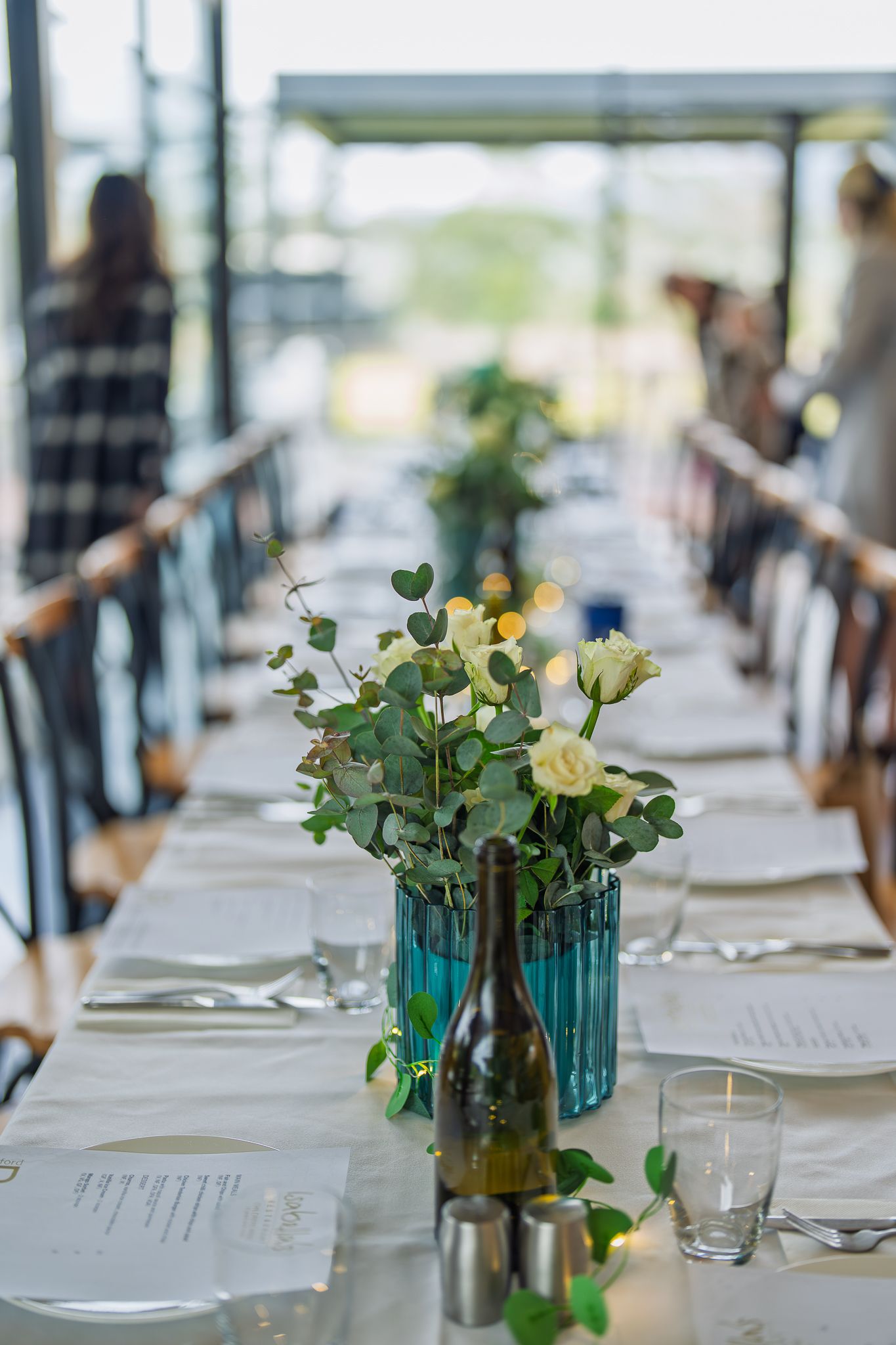 Elegant wedding reception table set with white roses in blue vases, candles, and wine bottles.
