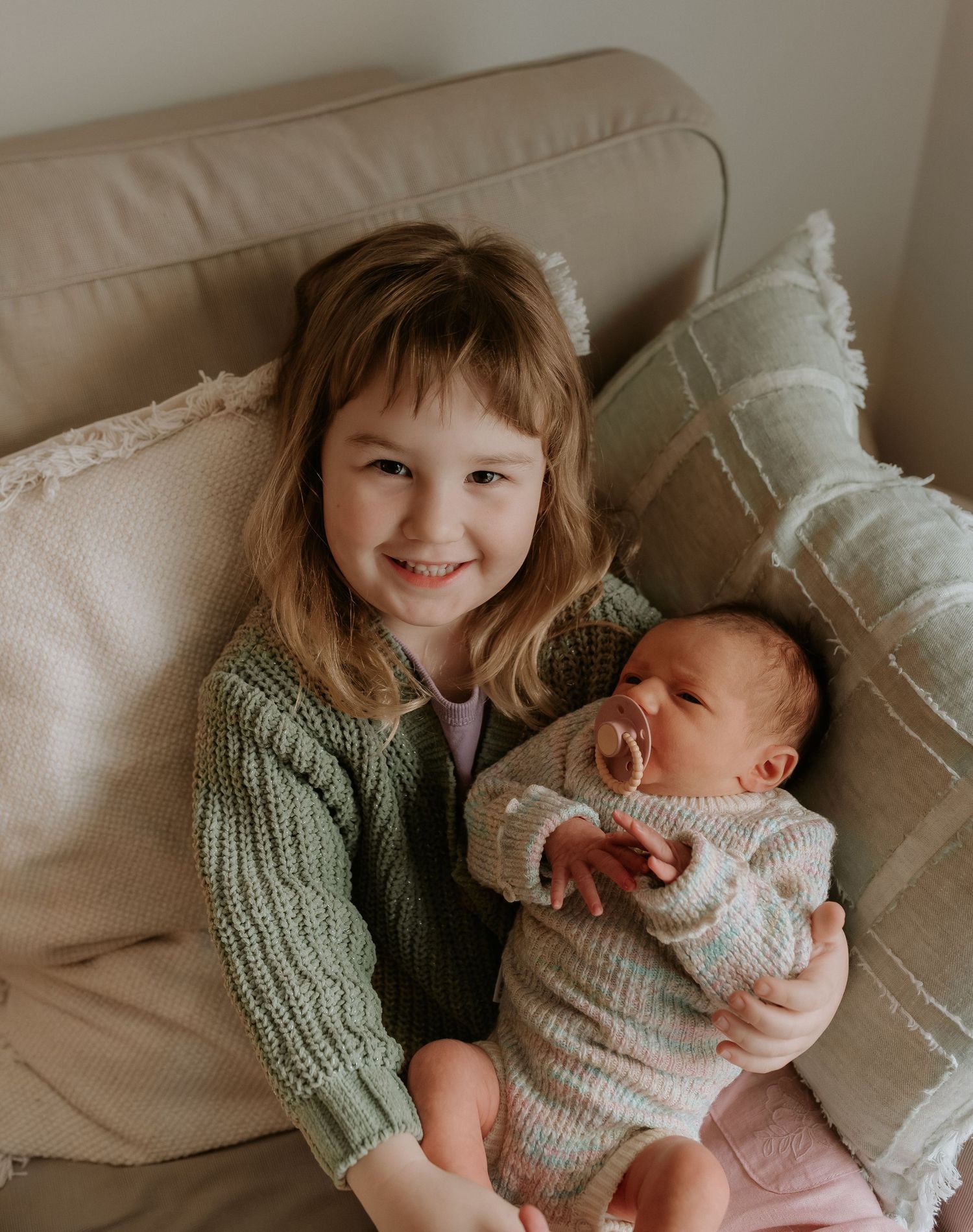 A tender moment between siblings as they cuddle together on a beige couch in matching knit sweaters.