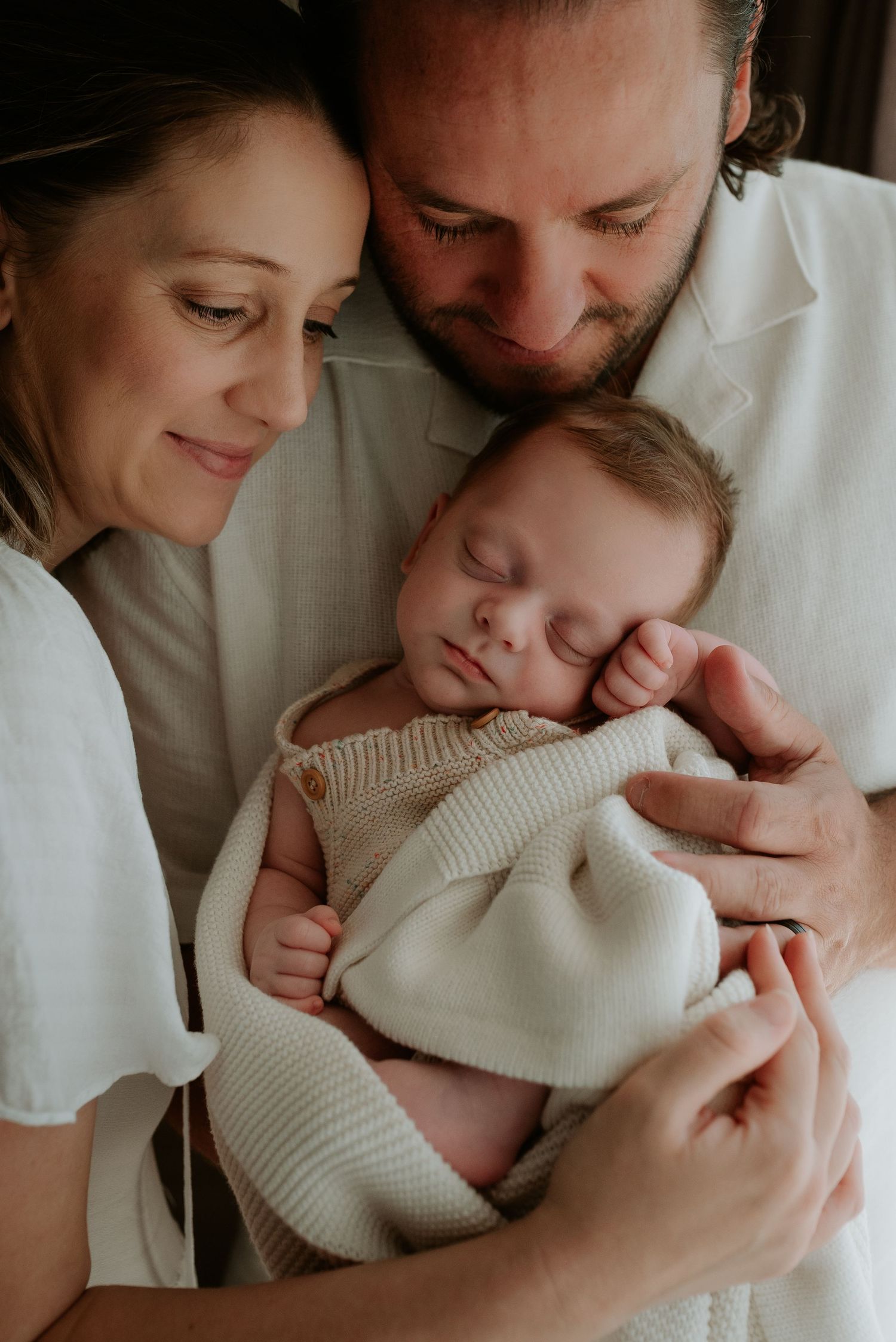 Intimate family portrait series showing tender moments with a sleeping newborn in cream knit clothing.