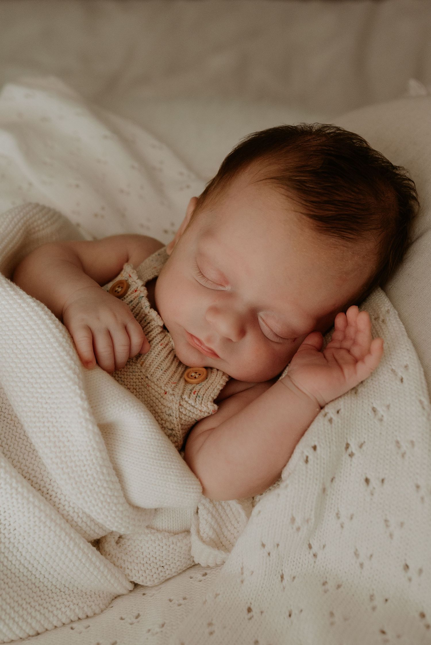 Serene newborn photography showing baby sleeping in cream knit clothing on white textured bedding.