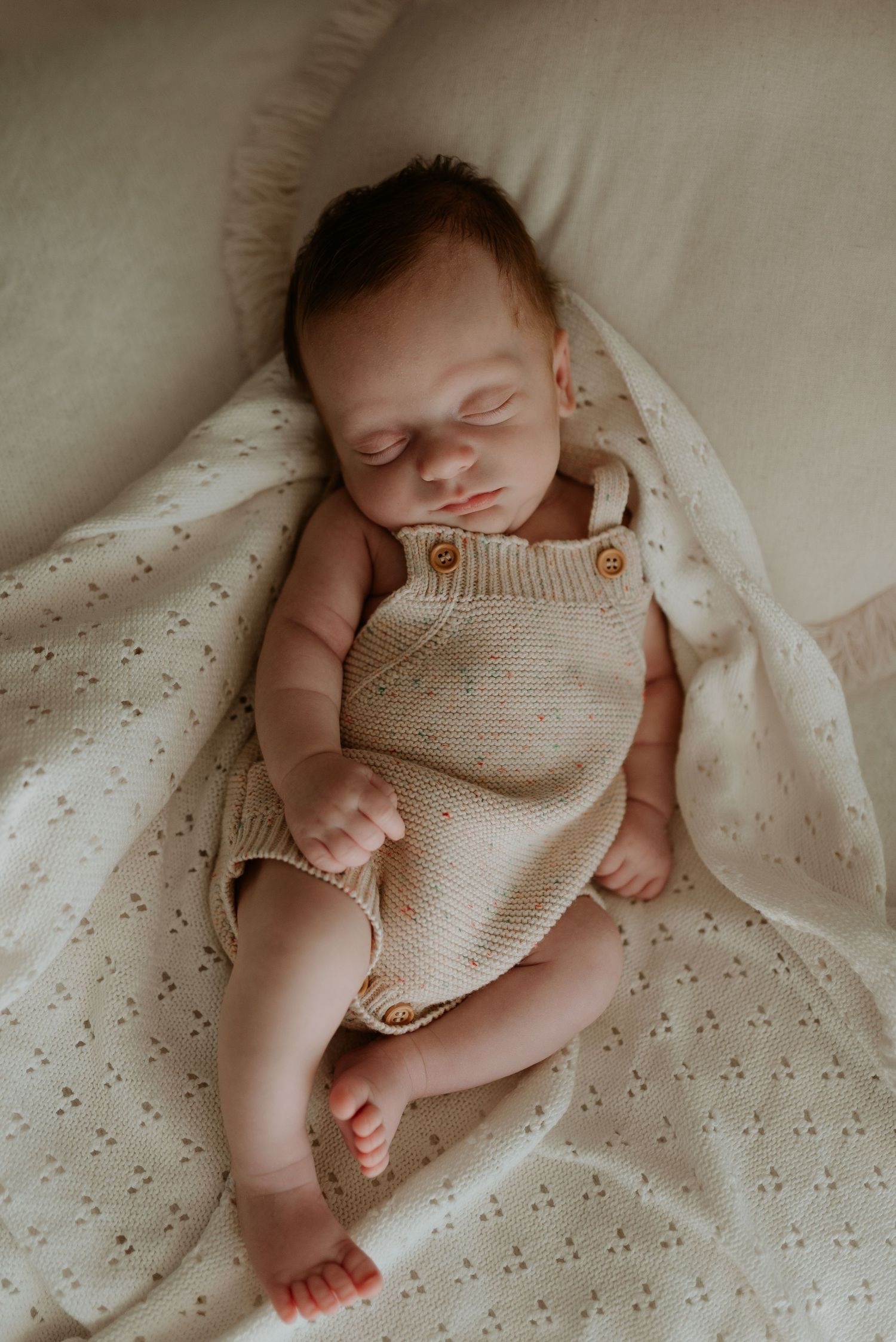 Artistic newborn portrait showing sleeping baby in beige knit outfit on white bedding.