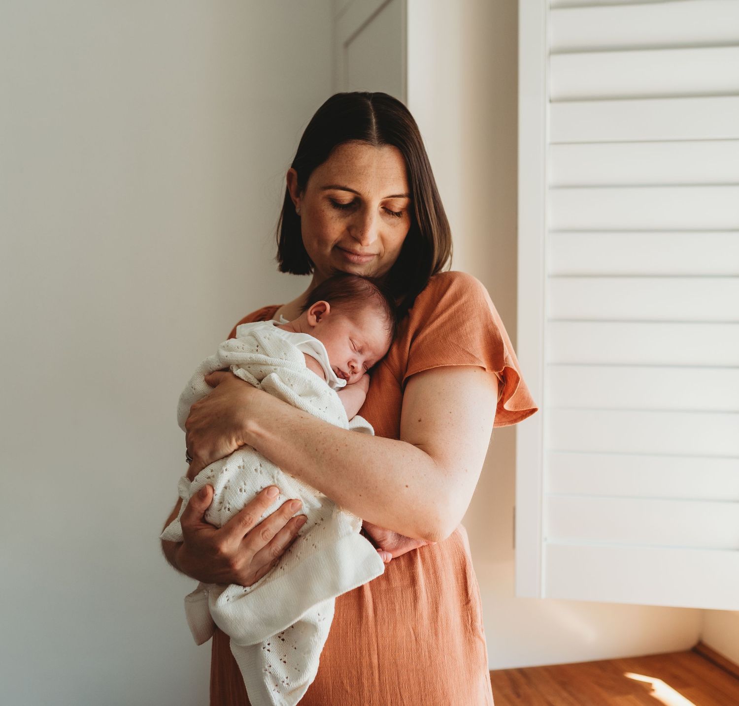 Mother in coral dress holding and bonding with newborn baby in bright room.