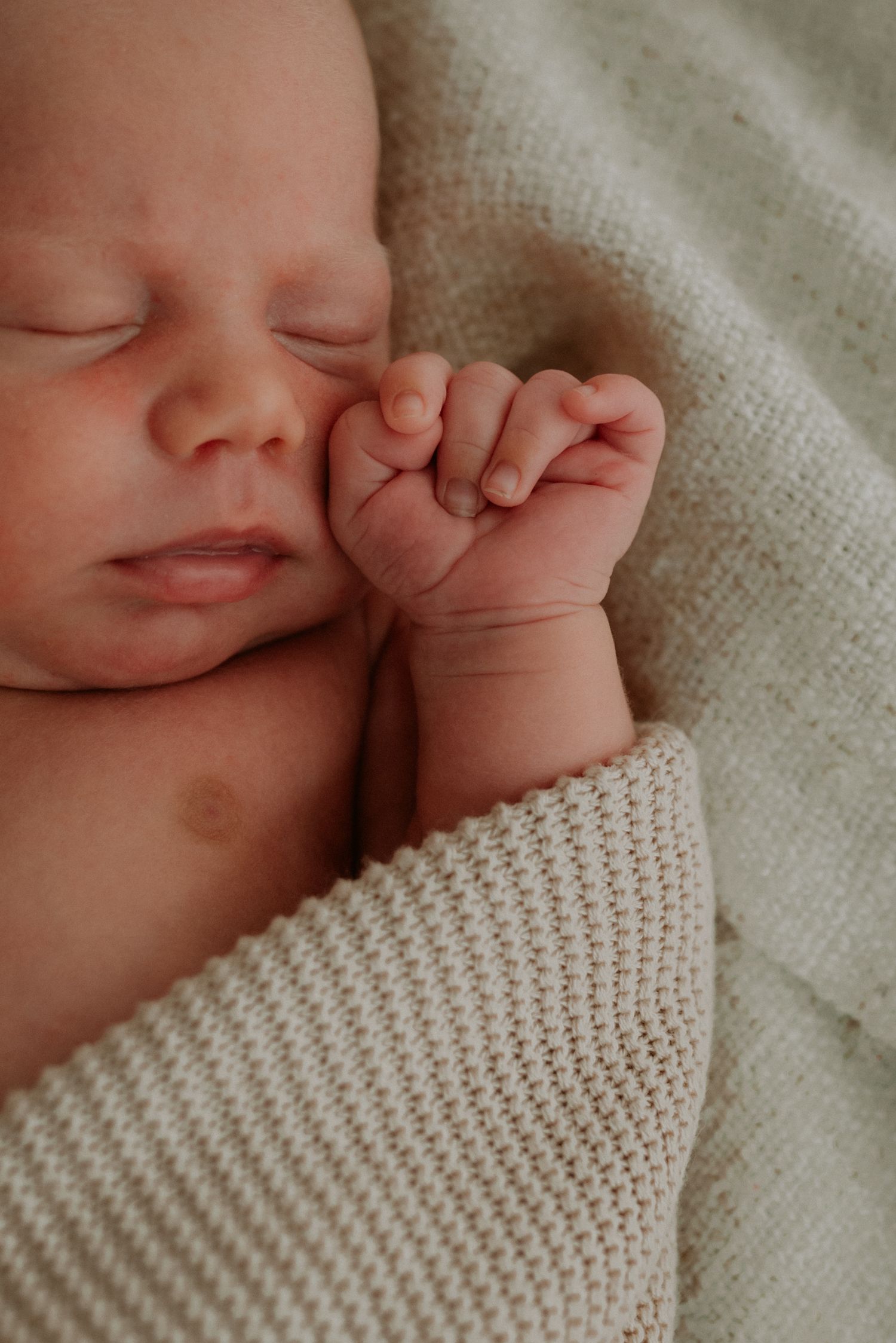 Sleeping newborn close-up with tiny hand near face on textured blanket.