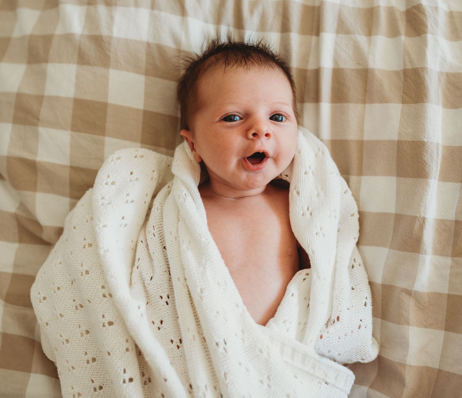 Close-up of newborn baby wrapped in white eyelet blanket against tan gingham background.