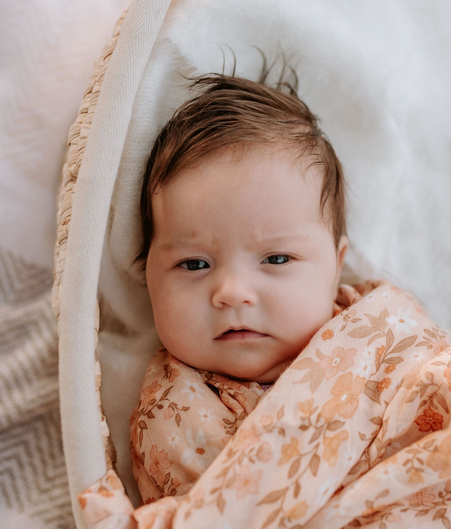 Baby wrapped in peach-colored floral swaddle lying on white bedding.