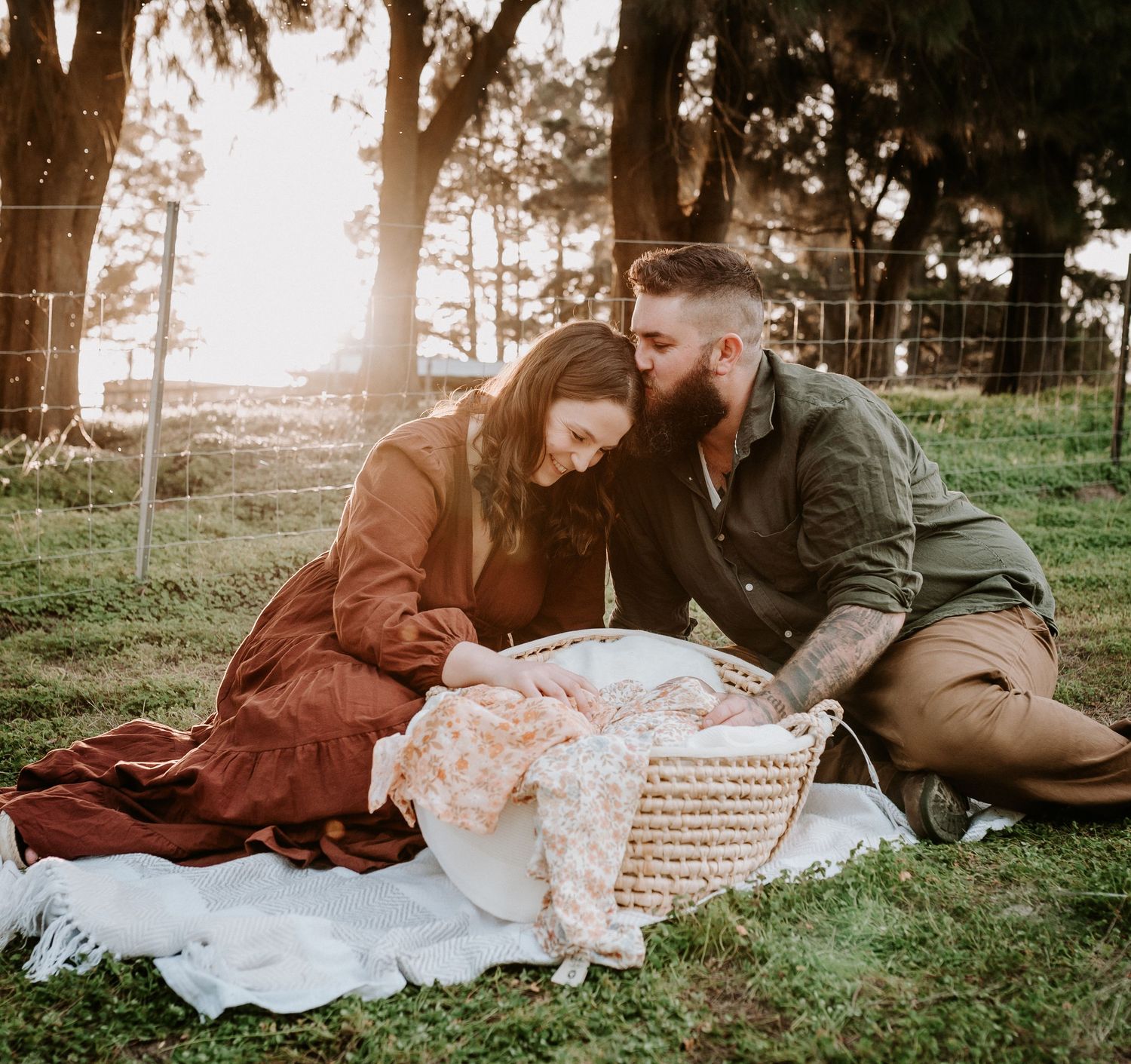 Family picnic scene at sunset with blanket spread on grass near trees.