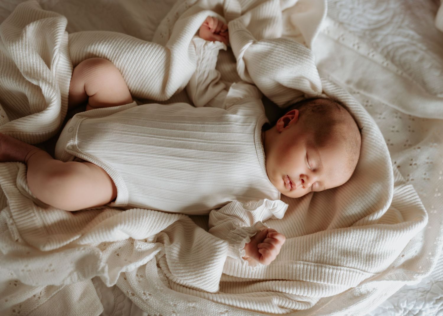 Baby in white knit clothing lying on textured cream bedding.