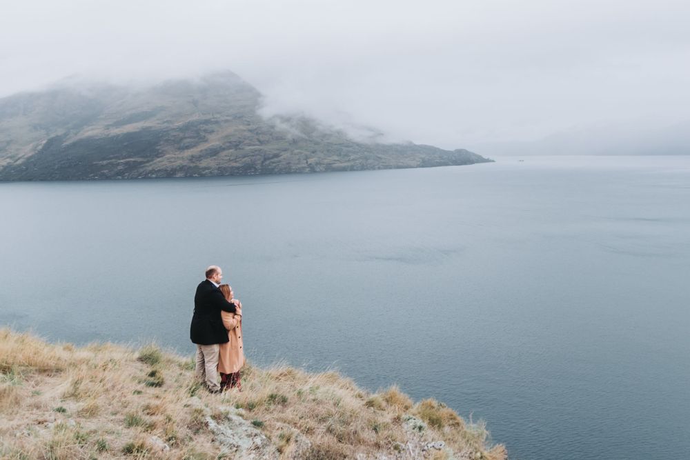 Two people embrace while enjoying panoramic views of a misty lake and mountains.
