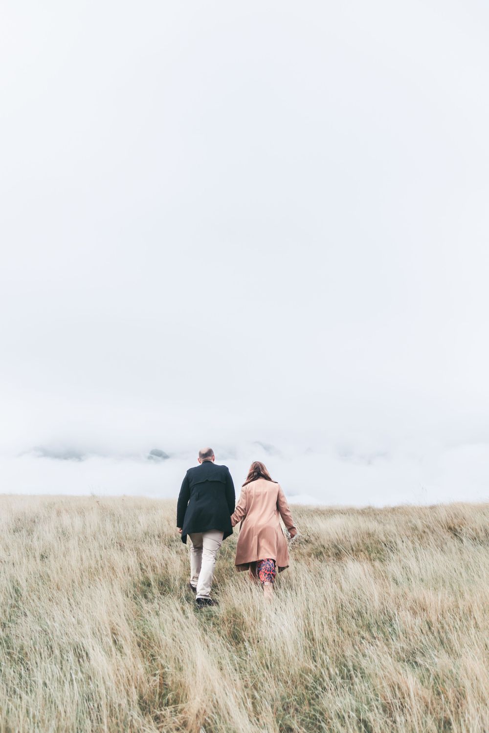 A couple walks through tall grass on a misty hillside path.