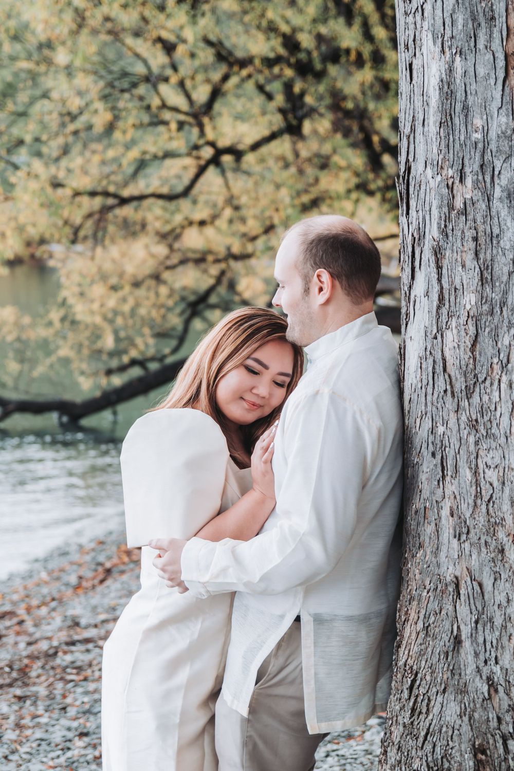 A romantic moment shared between two people leaning against a tree by the water.