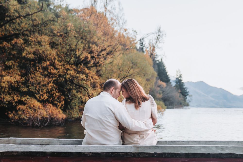 A couple sits close together on a bench overlooking a scenic autumn lake view.