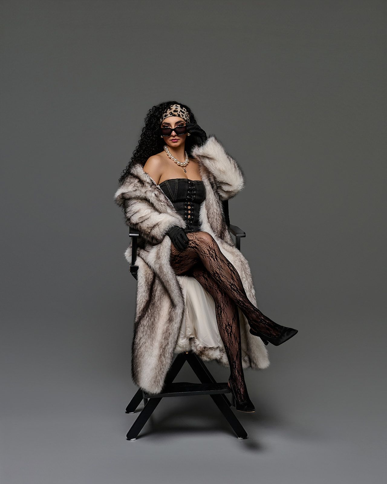 A figure in a luxurious fur coat and black boots poses dramatically on a chair against a gray studio backdrop.