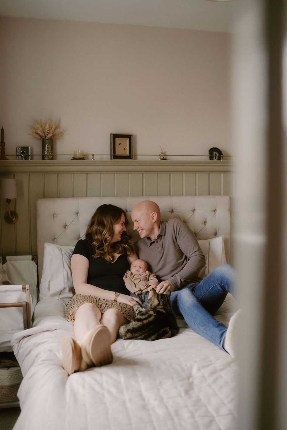 A young couple sat on a bed laughing toward eachother with their newborn in arms