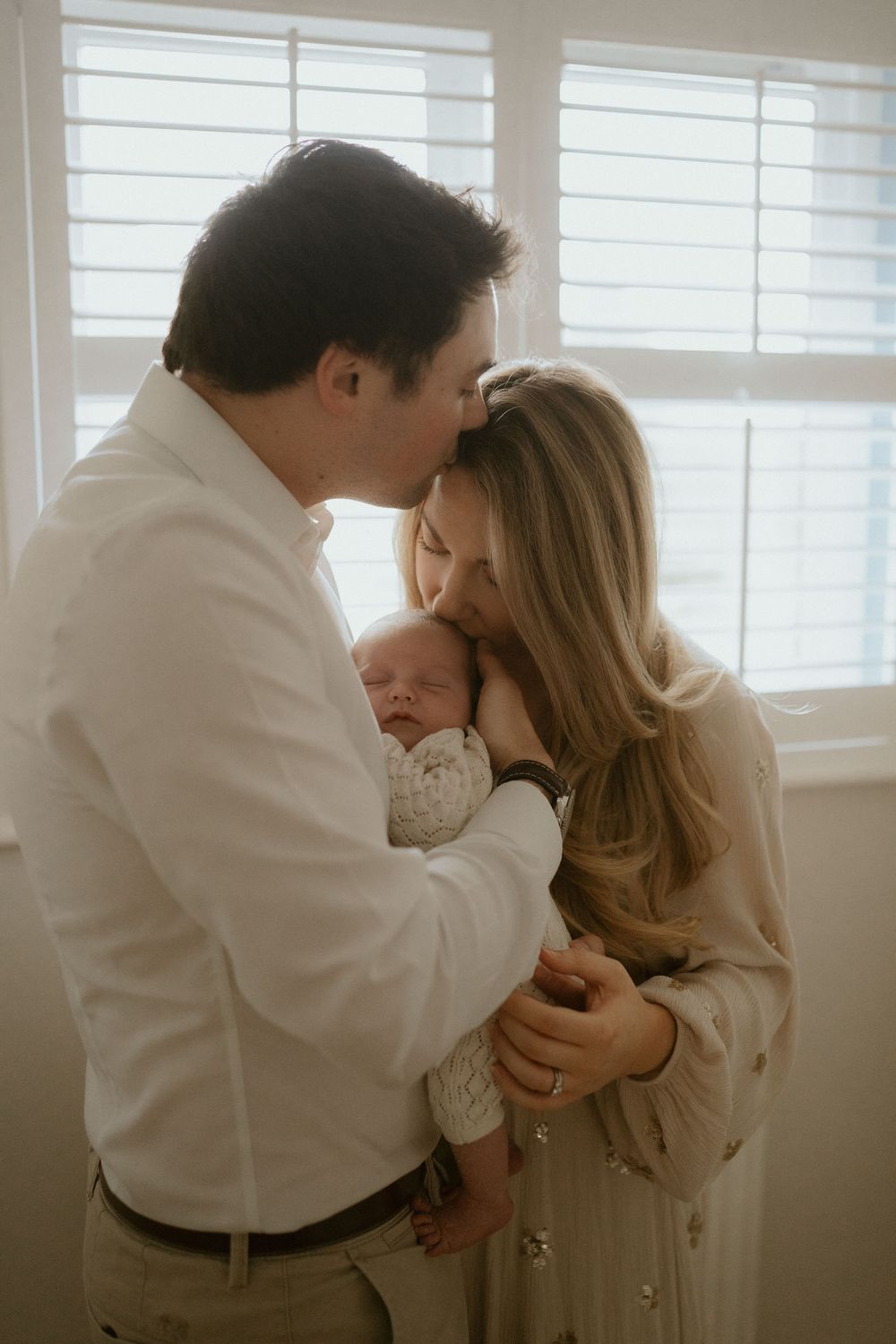 A father kissing the mother of his newborn child on her forehead whilst the mother kisses the babies back of the head whilst dad holds baby