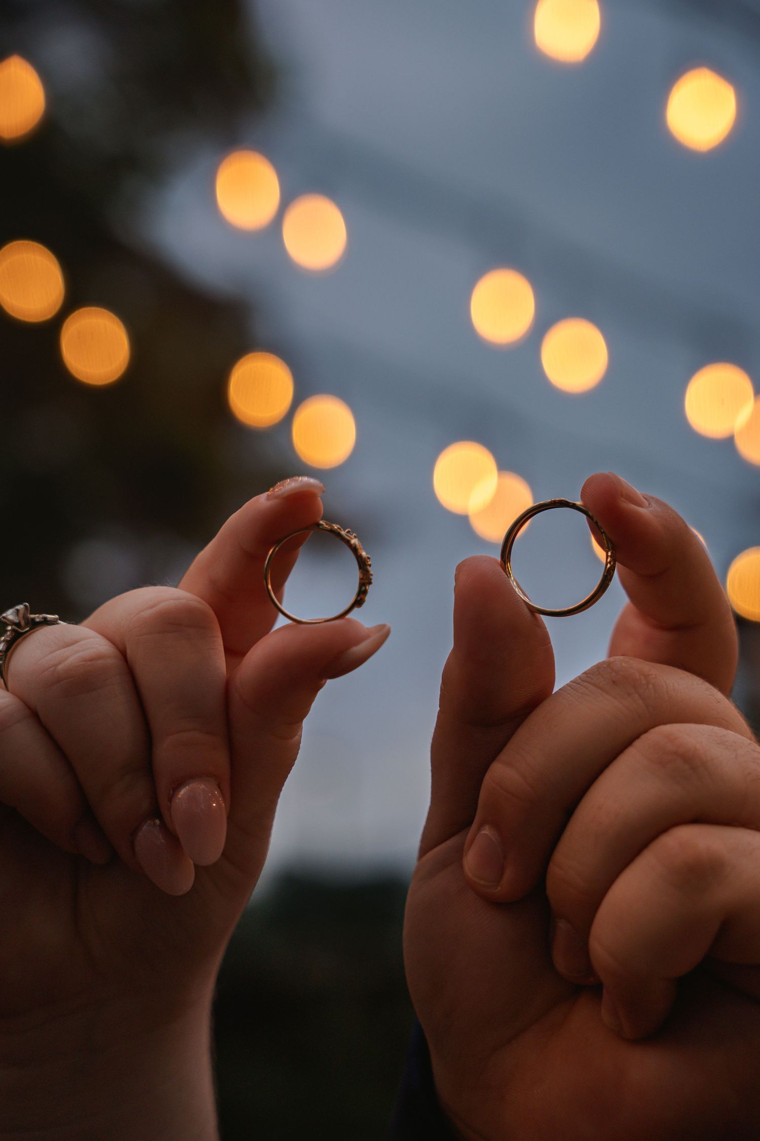Two hands hold wedding rings against a backdrop of glowing bokeh lights at dusk.