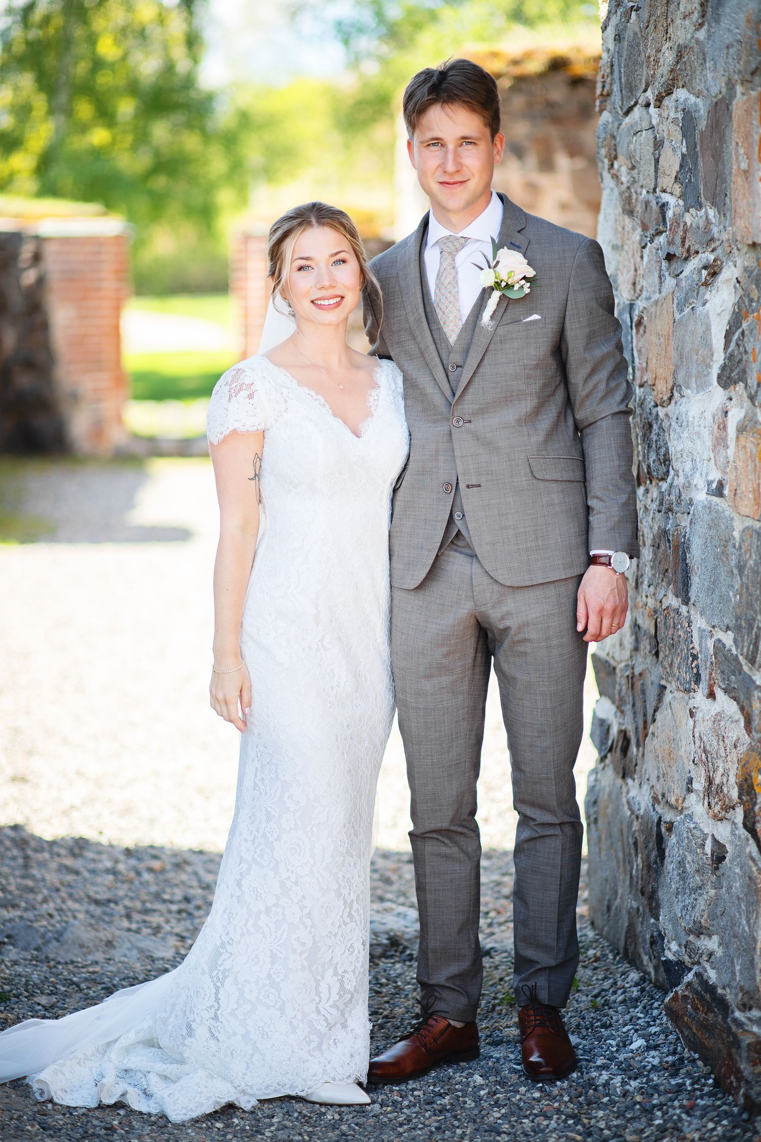 Bride in lace wedding dress and groom in grey suit posing beside stone wall outdoors.