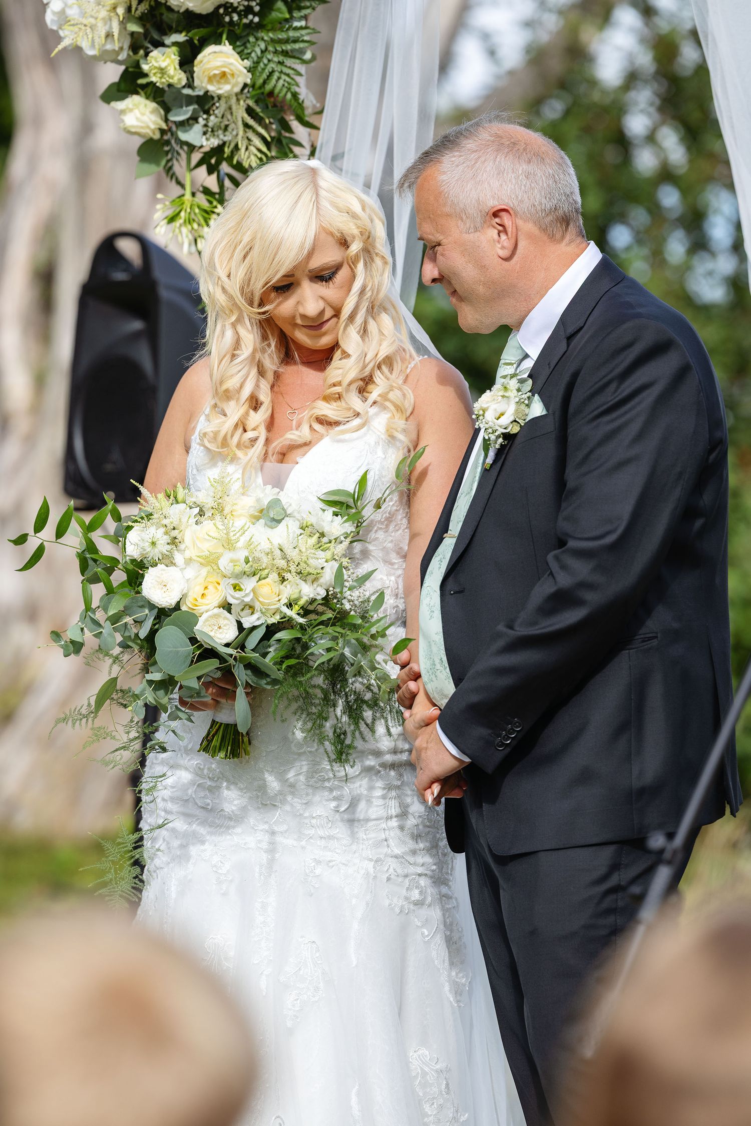 Bride in lace wedding dress and groom in grey suit posing beside stone wall outdoors.