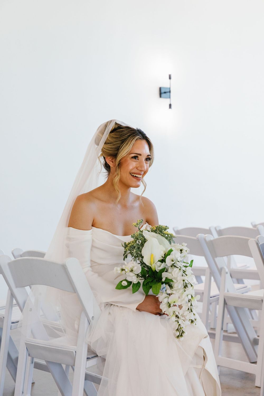 Wedding portrait of a smiling bride holding a cascading bouquet while seated in ceremony chairs.