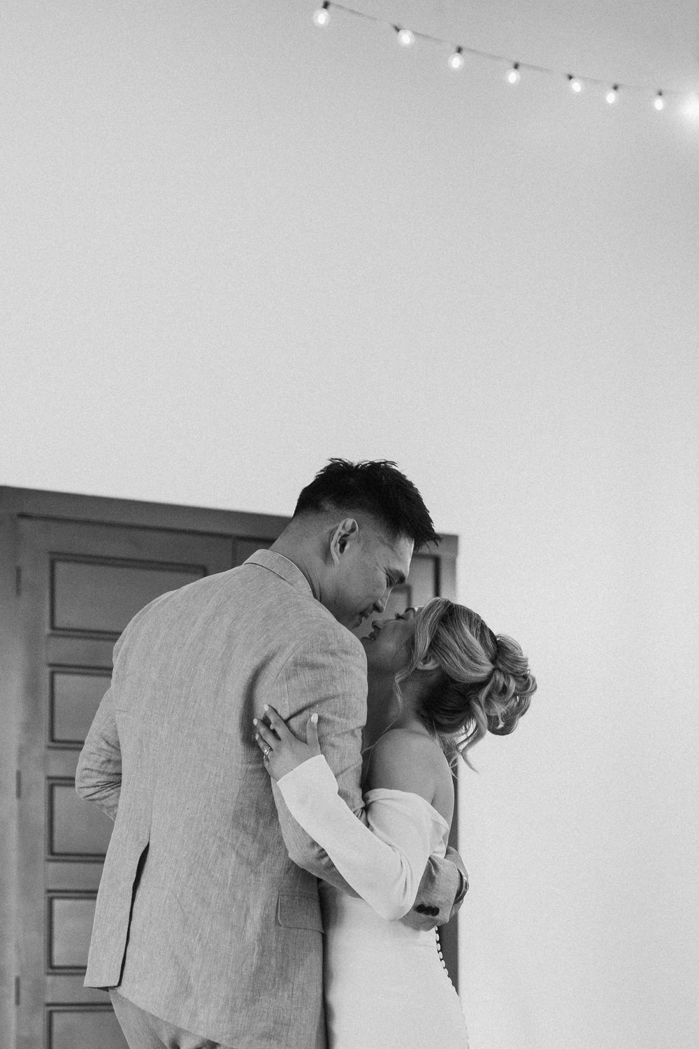 Black and white wedding photography of a bride and groom embracing and kissing under indoor string lights.